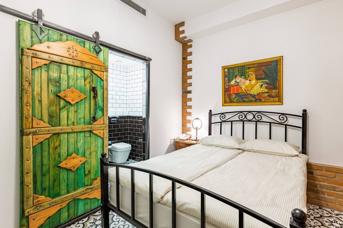 Ein Zimmer mit einem Bett, einer grünen Tür, einem Gemälde und einem kleinen Tisch mit einer Lampe.