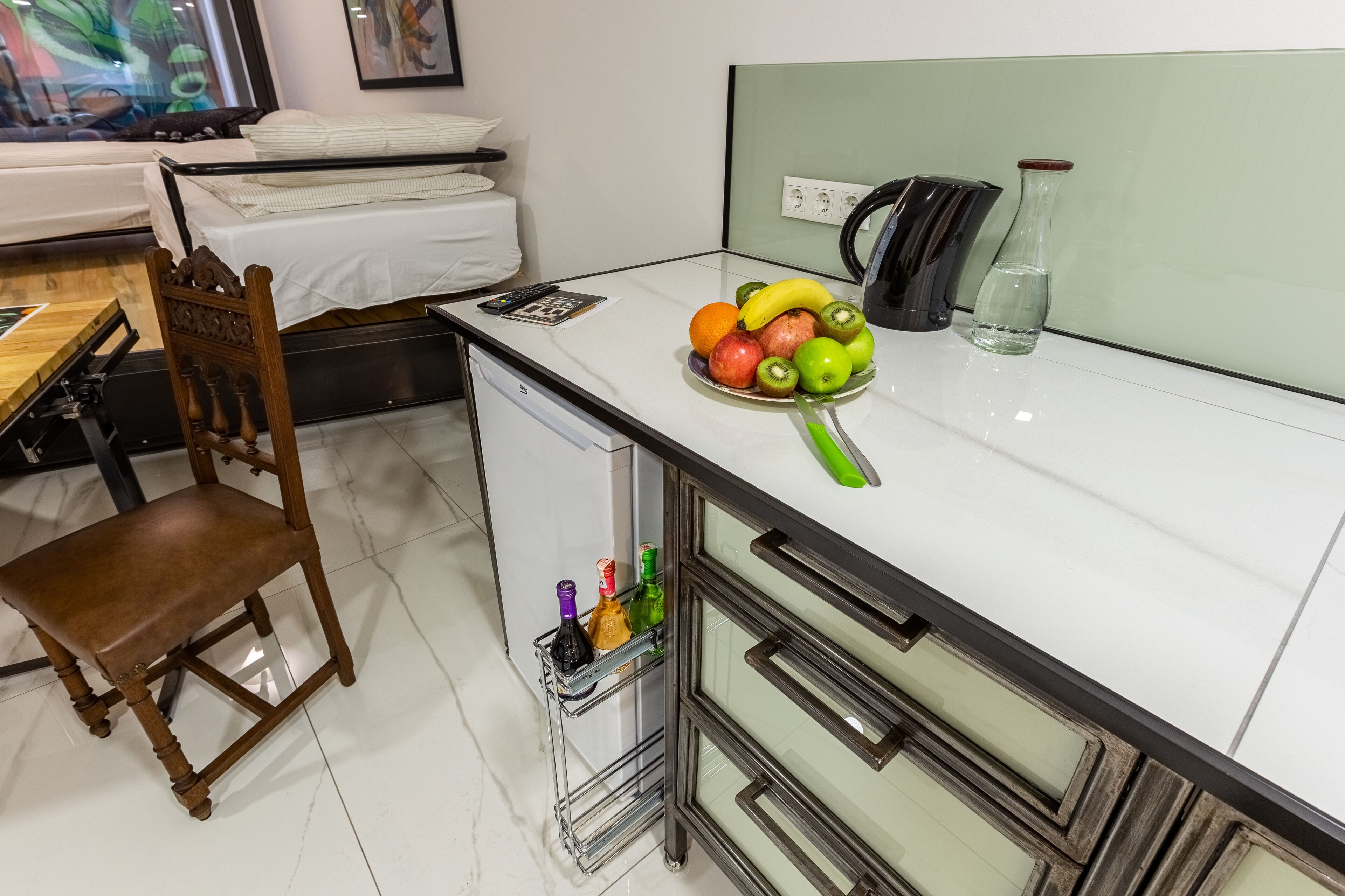 A compact kitchen with a white countertop, a wooden chair, a small fridge, and a fruit bowl with bananas, apples, and kiwi.
