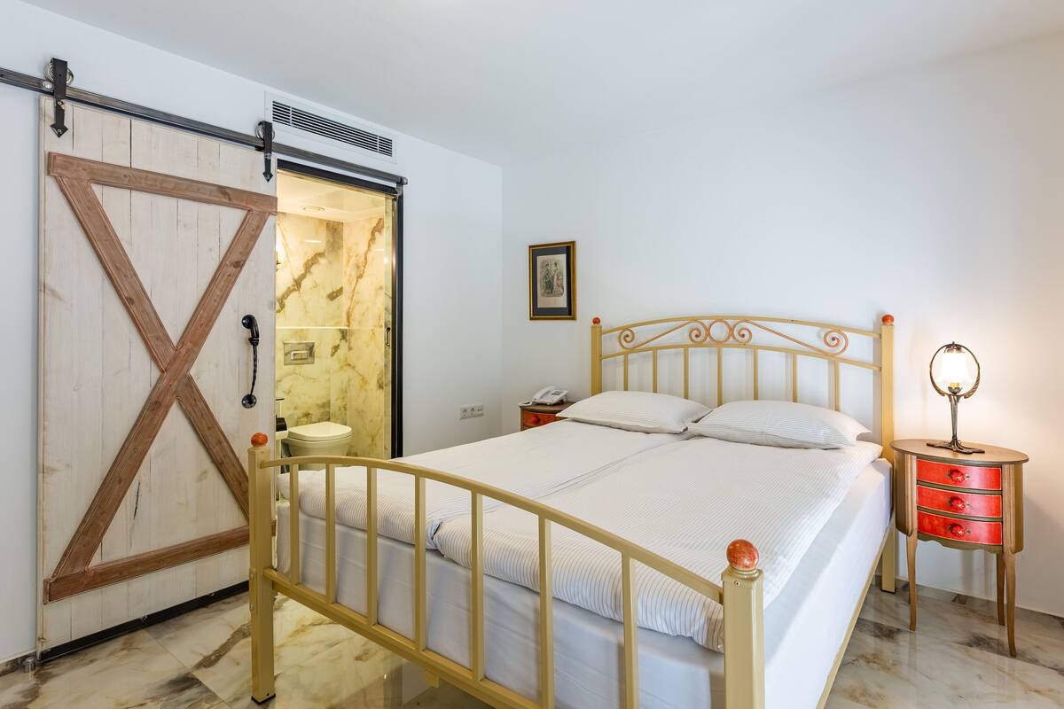 Ein Schlafzimmer mit einem Bett, einem Nachttisch mit Schubladen, einem kleinen Tisch, einem an der Wand befestigten Telefon, einem Bilderrahmen und einer Schiebetür mit Holzrahmen.