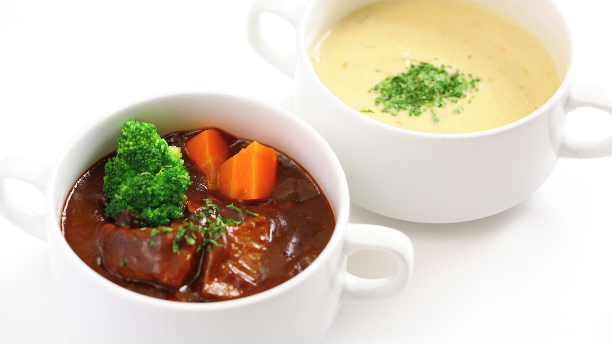Dos tazones de cerámica blanca, uno con un guiso oscuro y verduras, y el otro una crema con hierbas como decoración.