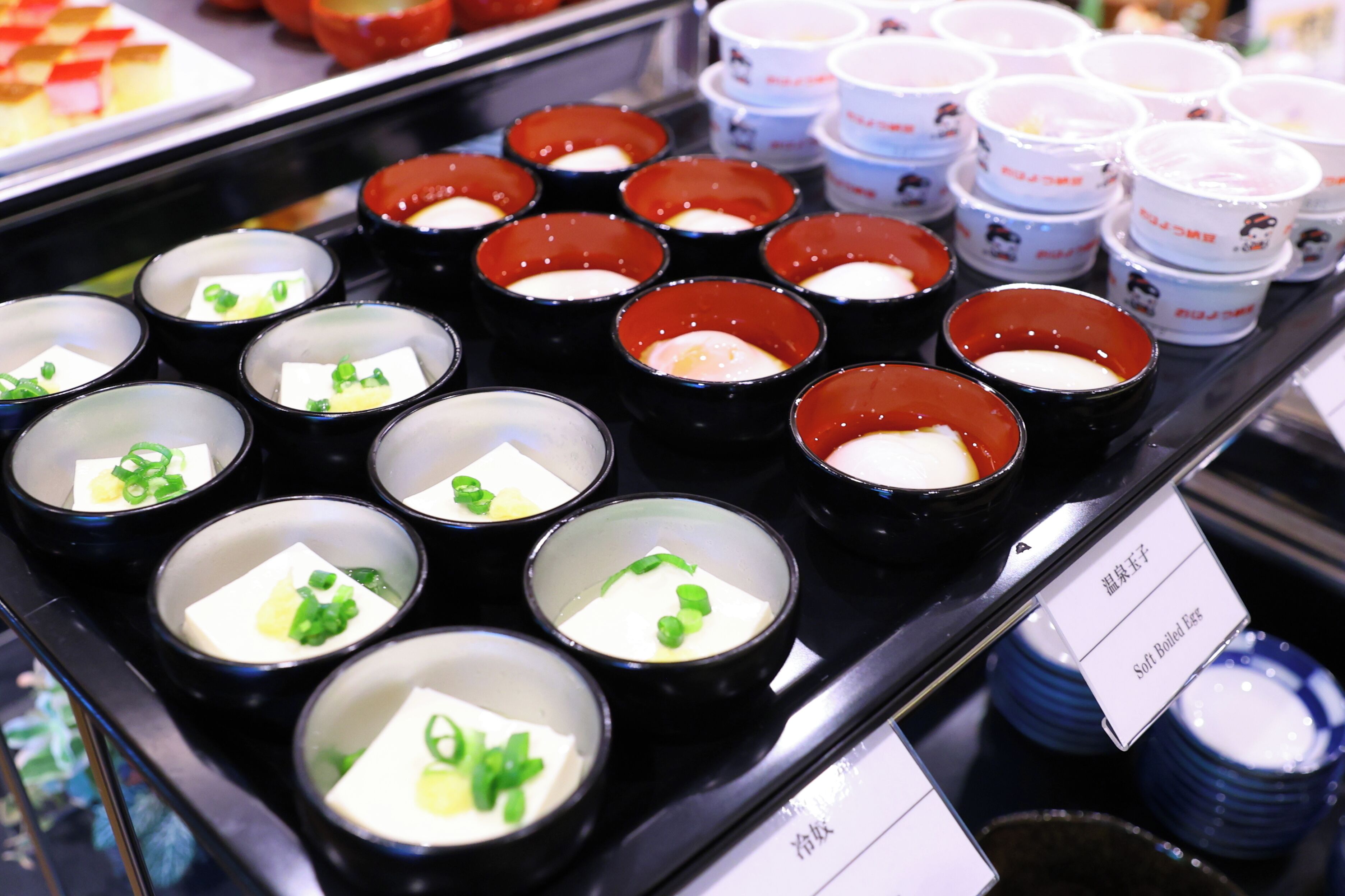 A display of various food items in small bowls, including tofu and soft-boiled eggs, arranged on a tray.