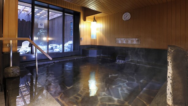 A hot spring bath area with a wooden interior, a clock on the wall, and a view of the outside through large glass doors.