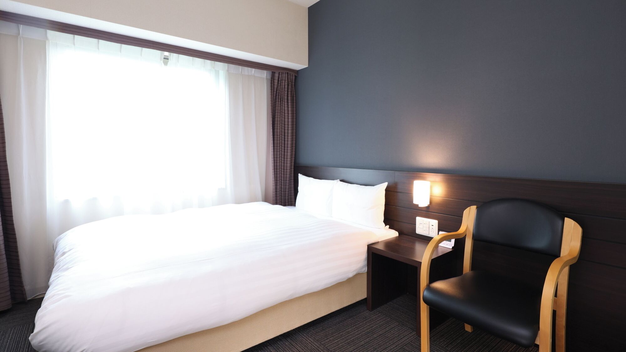 Habitación de hotel con cama, mesita de noche, una silla y una lámpara fijada a la pared.