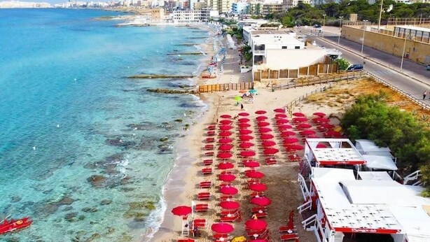 A beach with rows of red umbrellas and white lounge chairs, a clear blue sea, and buildings in the background. - La Casa del Turista (Gallipoli)