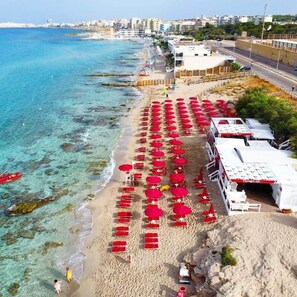A beach with rows of red umbrellas and white lounge chairs, a clear blue sea, and buildings in the background. - La Casa del Turista (Gallipoli)