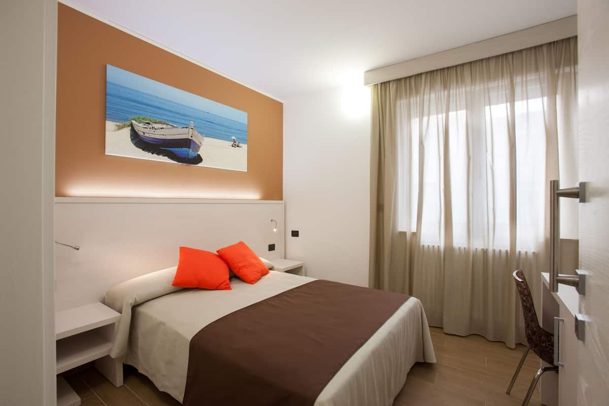 A hotel room with a bed, a nightstand, a chair, a painting of a beach scene, and a window with curtains.