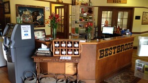 A reception desk with a wooden counter labeled "BESTBROOK," a computer, and a "Clydesdale Room" sign. There are jars of honey on the counter, a vase with flowers, and a fire extinguisher sign on the wall. - Bestbrook Mountain Resort (Tregony)