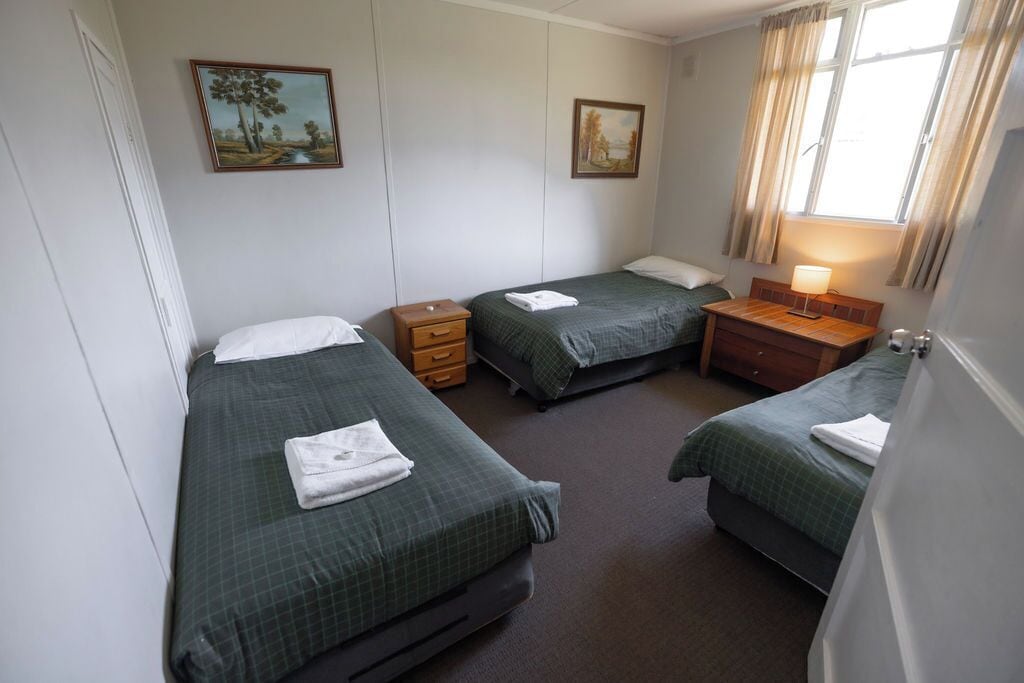 Een kamer met drie eenpersoonsbedden, elk met een groene sprei en witte kussens. Er zijn houten nachtkastjes met lampen, en de muren zijn versierd met twee landschapsschilderijen.