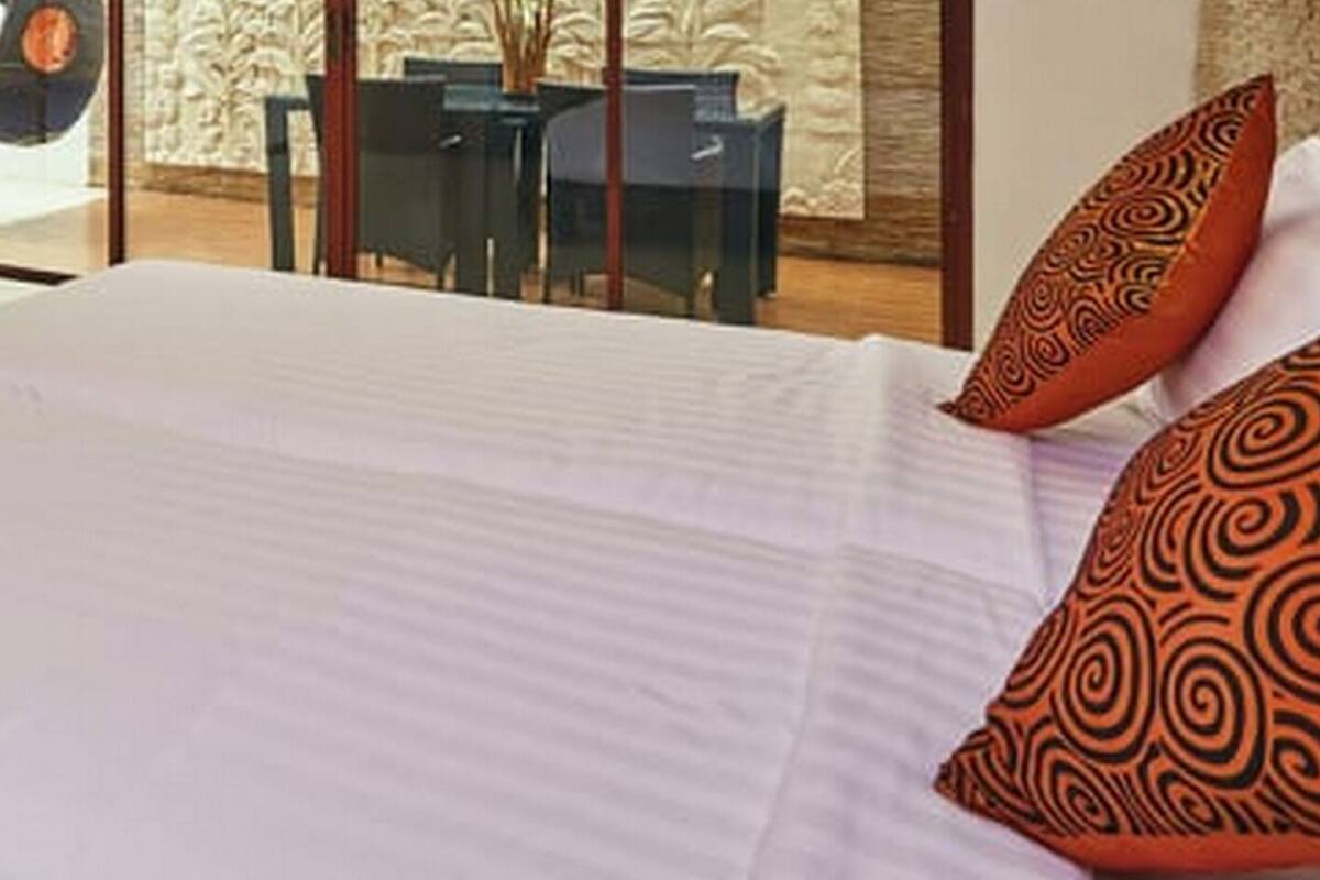 A hotel room with a large bed, a small table, and a chair. There is a view of a pool and a dining area through large windows.