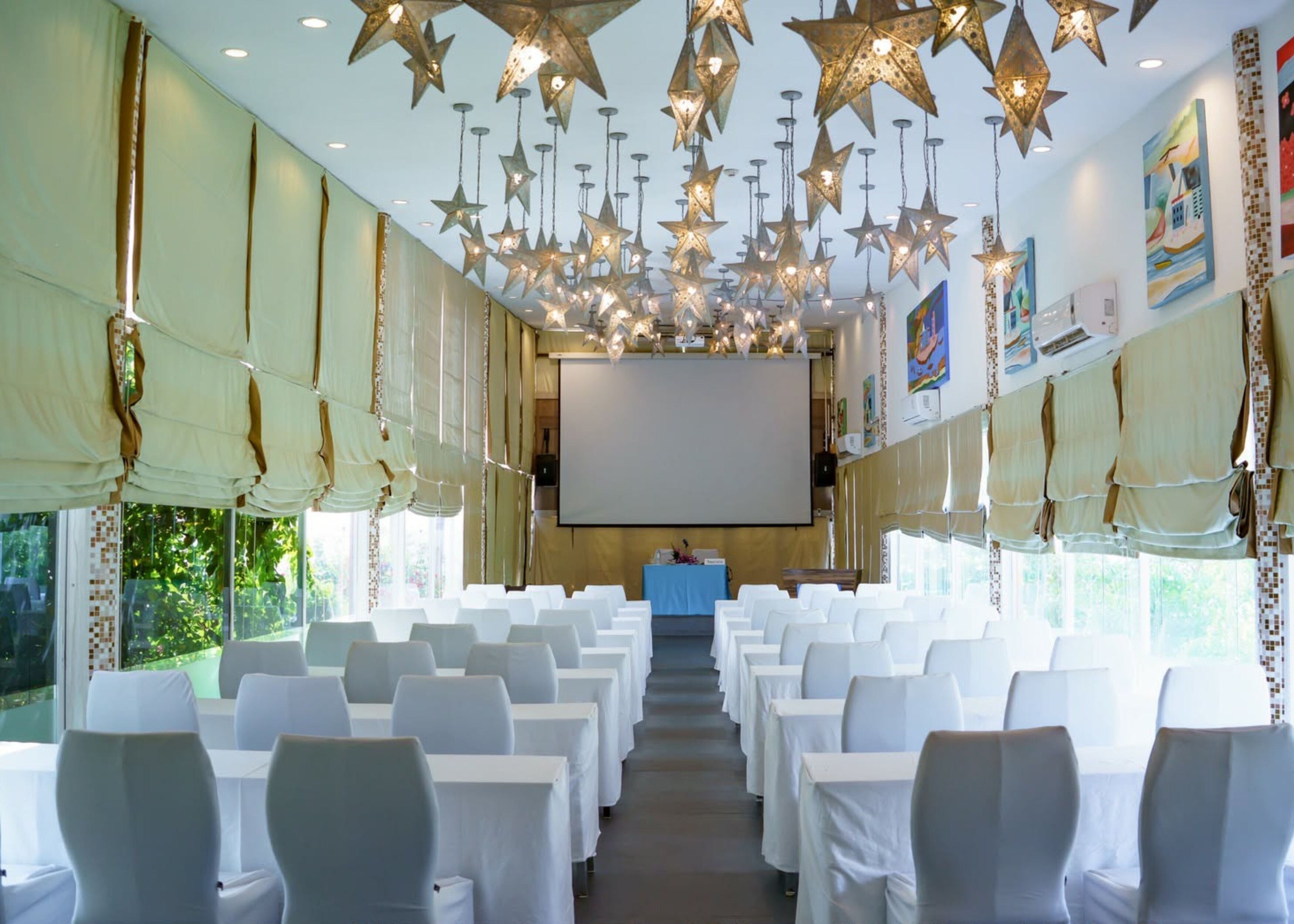Un salón de conferencias con sillas blancas, una pantalla de proyector y lámparas colgantes con forma de estrella.