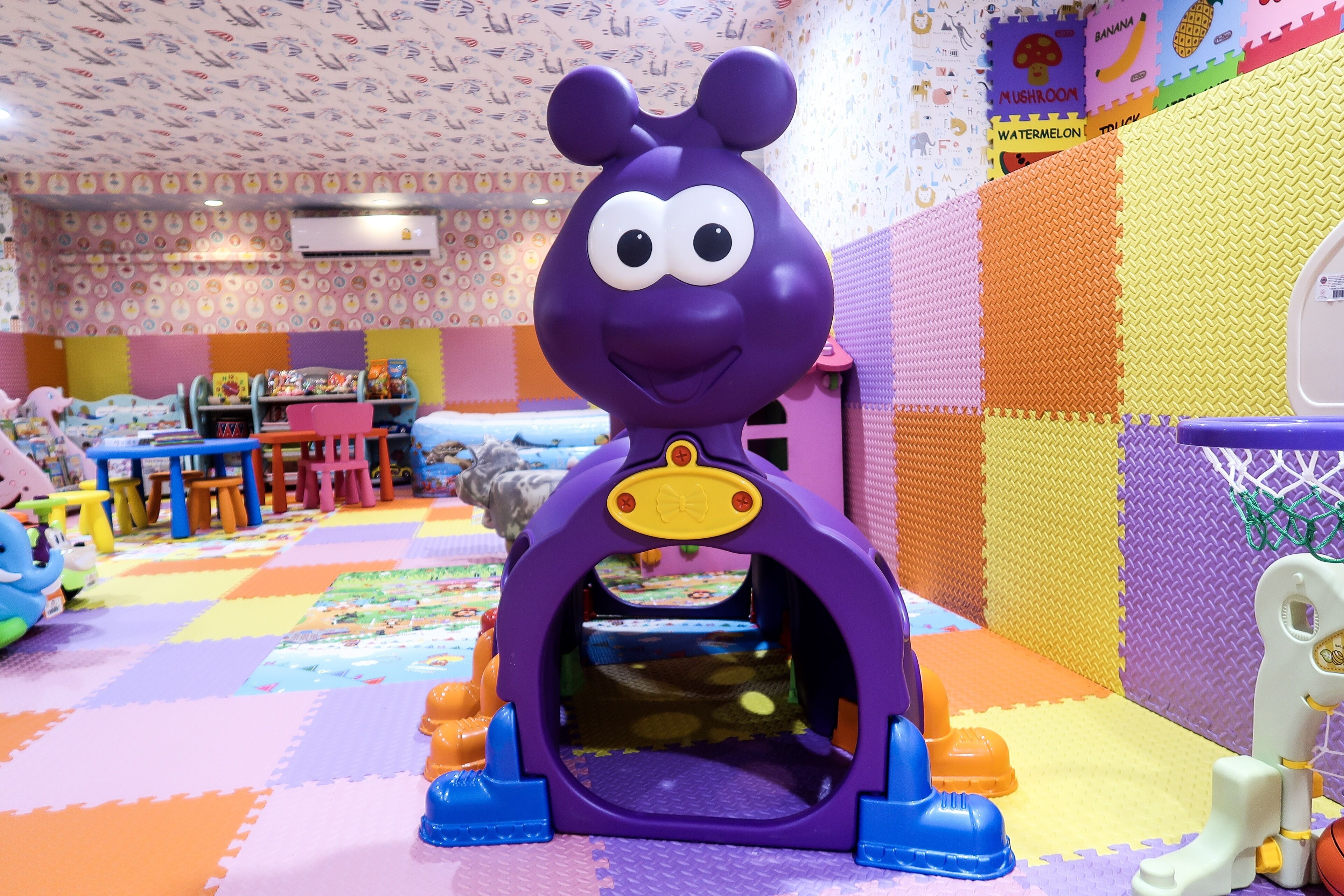 Un espace de jeux pour enfants avec une structure en plastique violet, des tapis en mousse de couleurs vives et un mur orné de posters éducatifs.