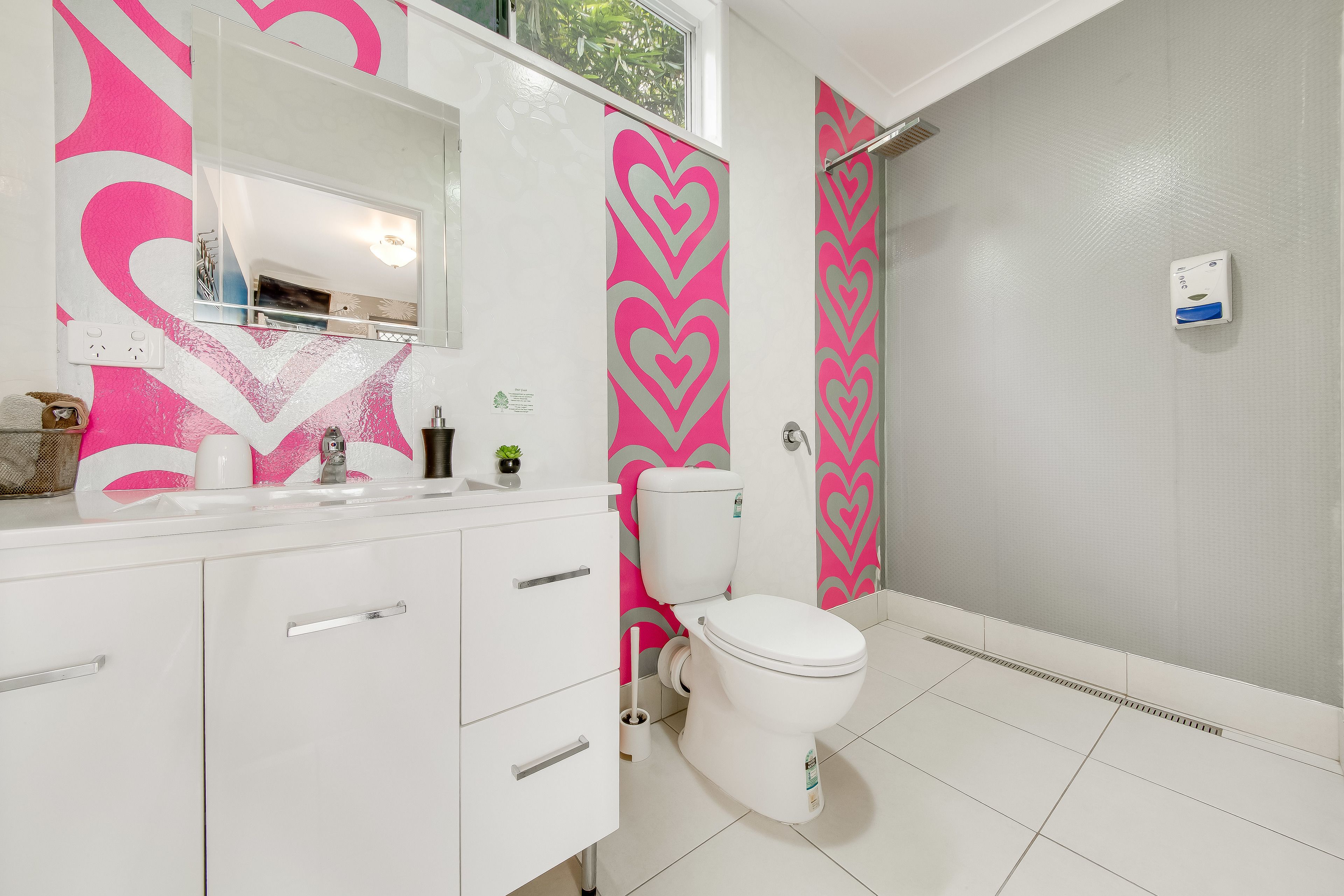 Un baño con lavamanos blanco, espejo e inodoro. Cuenta con una zona de ducha con puerta de vidrio y cortina blanca. Las paredes presentan un estampado de corazones rosa y blanco.