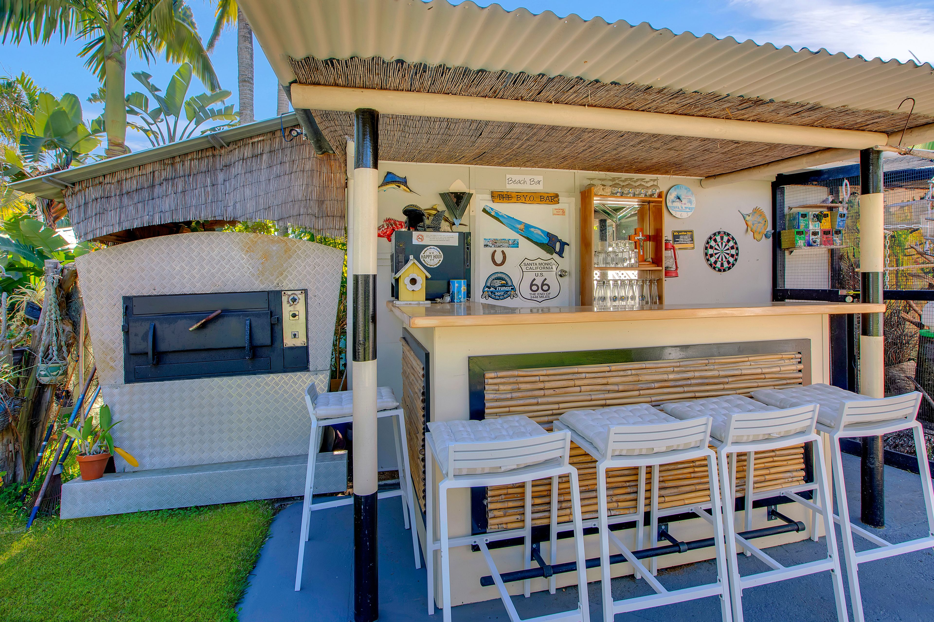 Eine Strandbar mit rustikalem Design, darunter eine Holztheke, weiße Barhocker und eine Wand, die mit Stranddekorationen gestaltet ist.