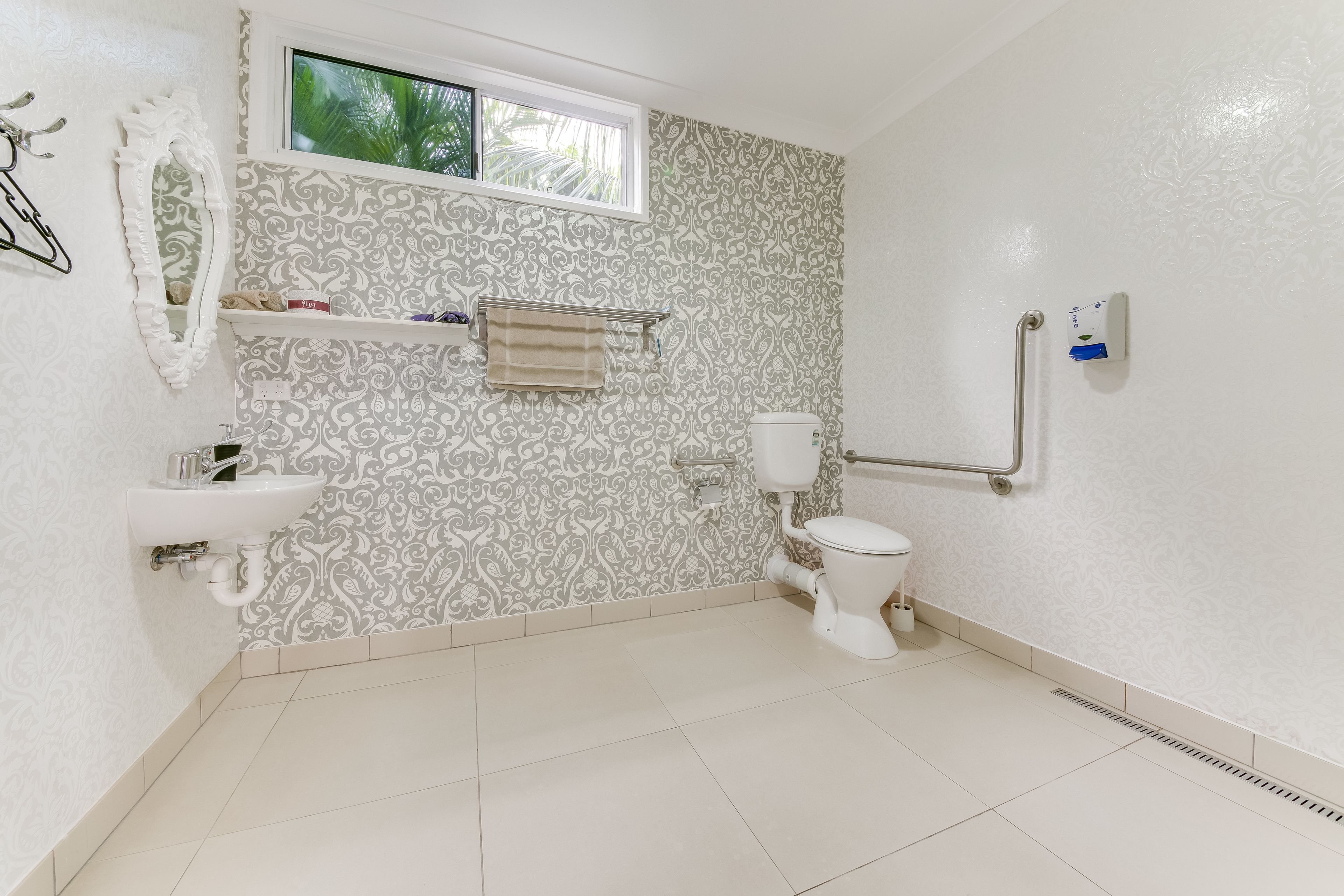 Une salle de bain avec un mur à motifs, une fenêtre, un lavabo, des toilettes et une barre d’appui.