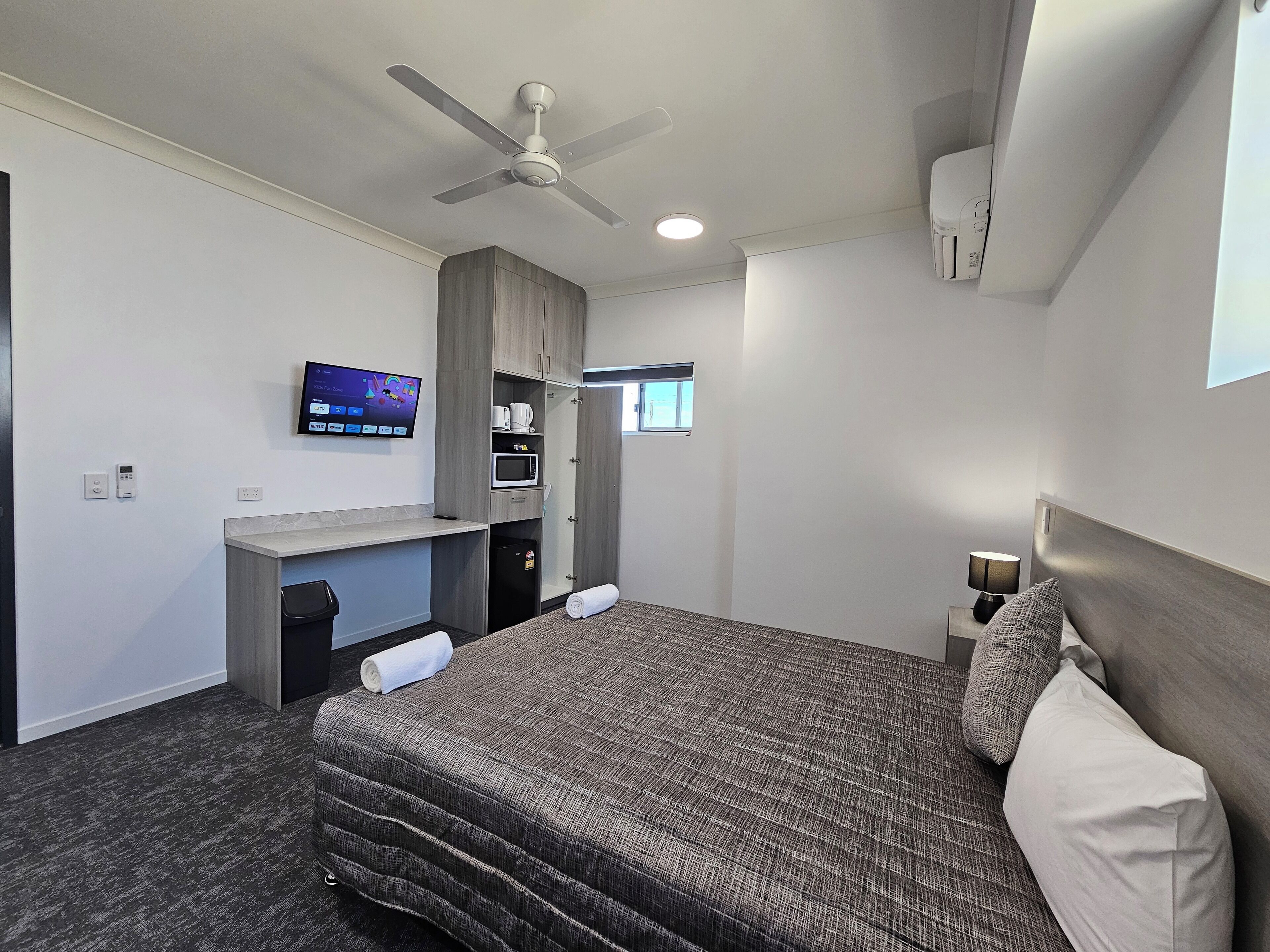 A modern hotel room with a bed, a desk, a television, a ceiling fan, and a small window.
