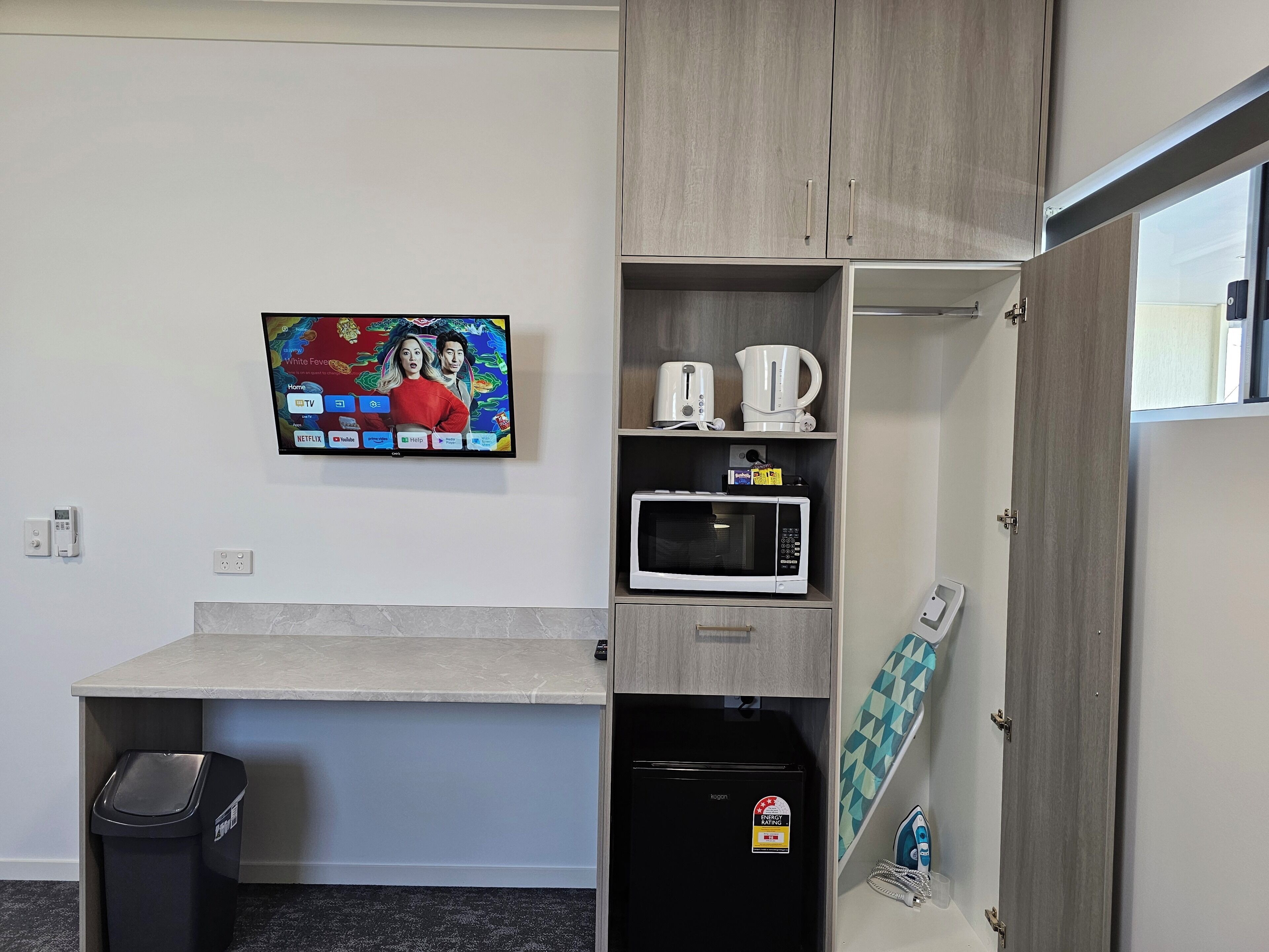 Una kitchenette compacta con microondas, hervidor y cafetera en estantes, un refrigerador pequeño y un televisor montado en la pared mostrando un programa.