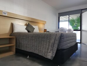 A modern hotel room with a bed, a wooden headboard, a nightstand, and a window with a view of palm trees.