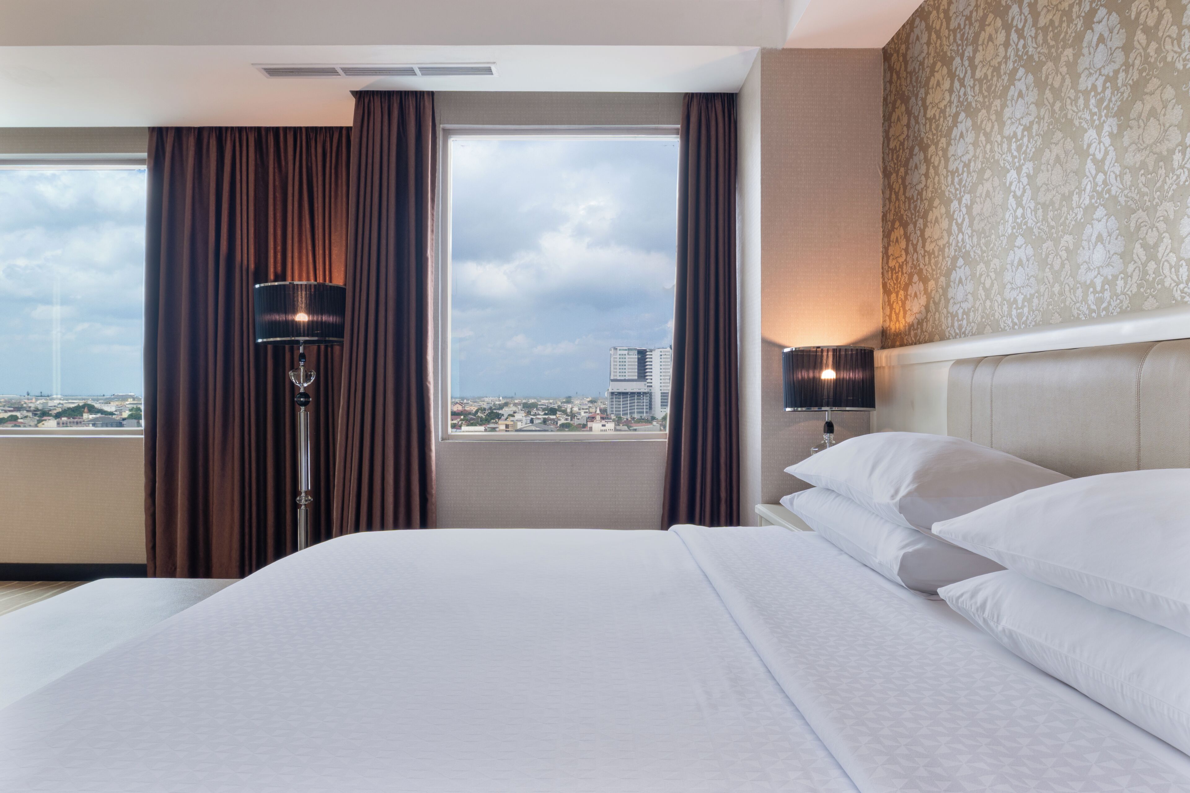 A hotel room with a large bed, a view of the city, a lamp, and a wall with a floral pattern.