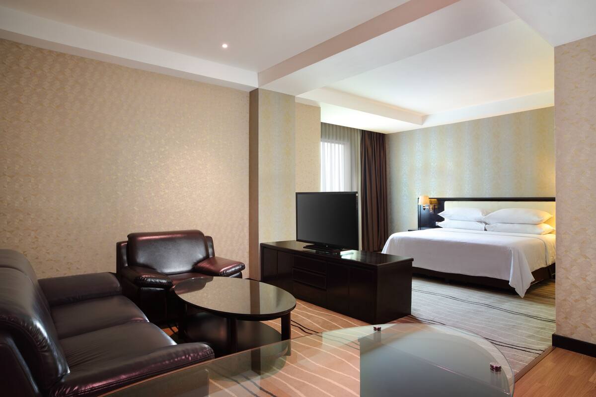 Habitación de hotel con una cama, televisor, sofá de cuero, mesita de centro de vidrio y un escritorio pequeño.
