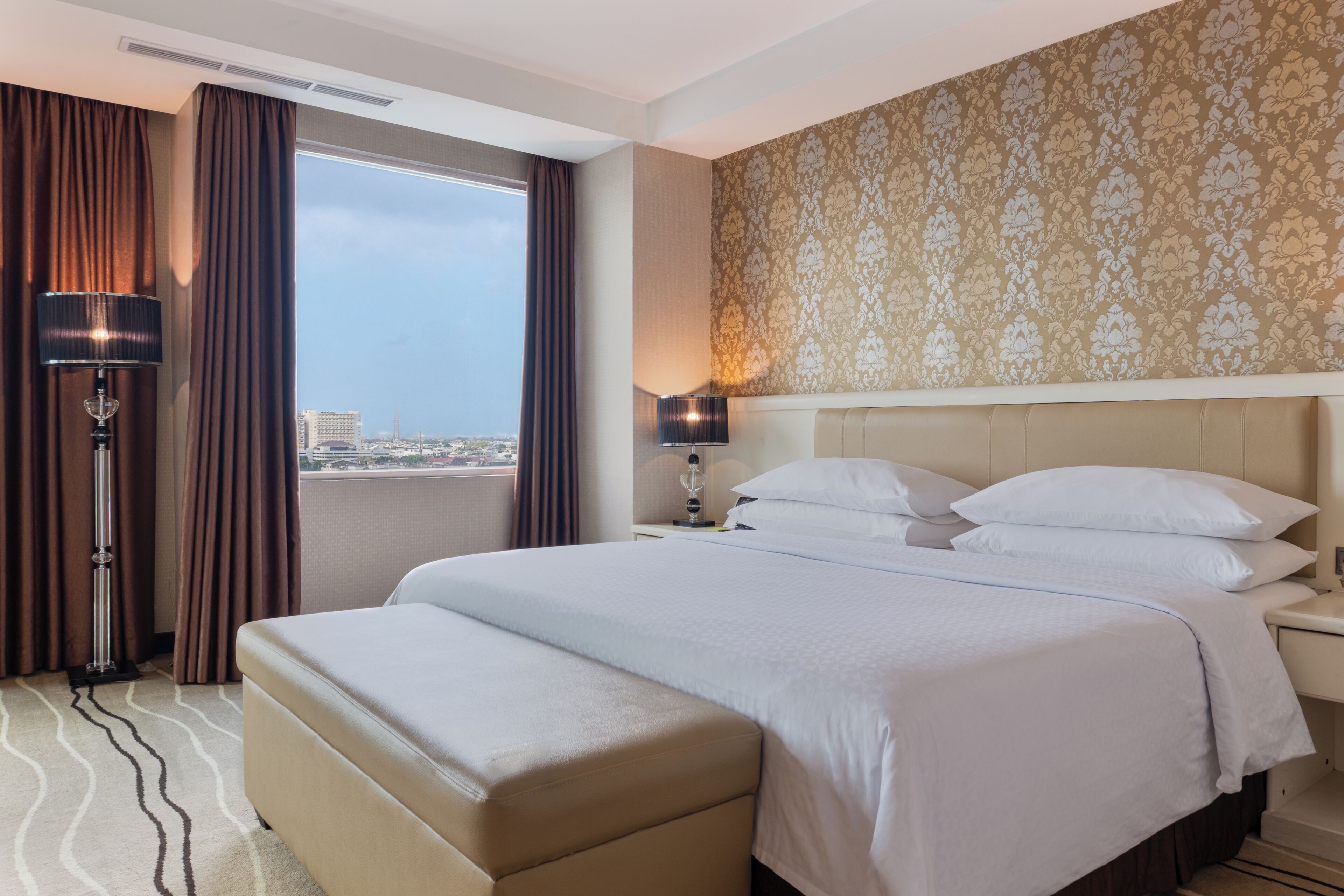 A hotel room with a large bed, a bedside table, a lamp, a window with curtains, and a patterned wall.