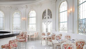 Un elegante comedor con mesas y sillas blancas, ventanas ornamentadas y una barra de mármol.