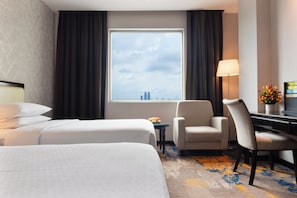 A hotel room with two beds, a desk, a chair, a lamp, and a view of the cityscape.