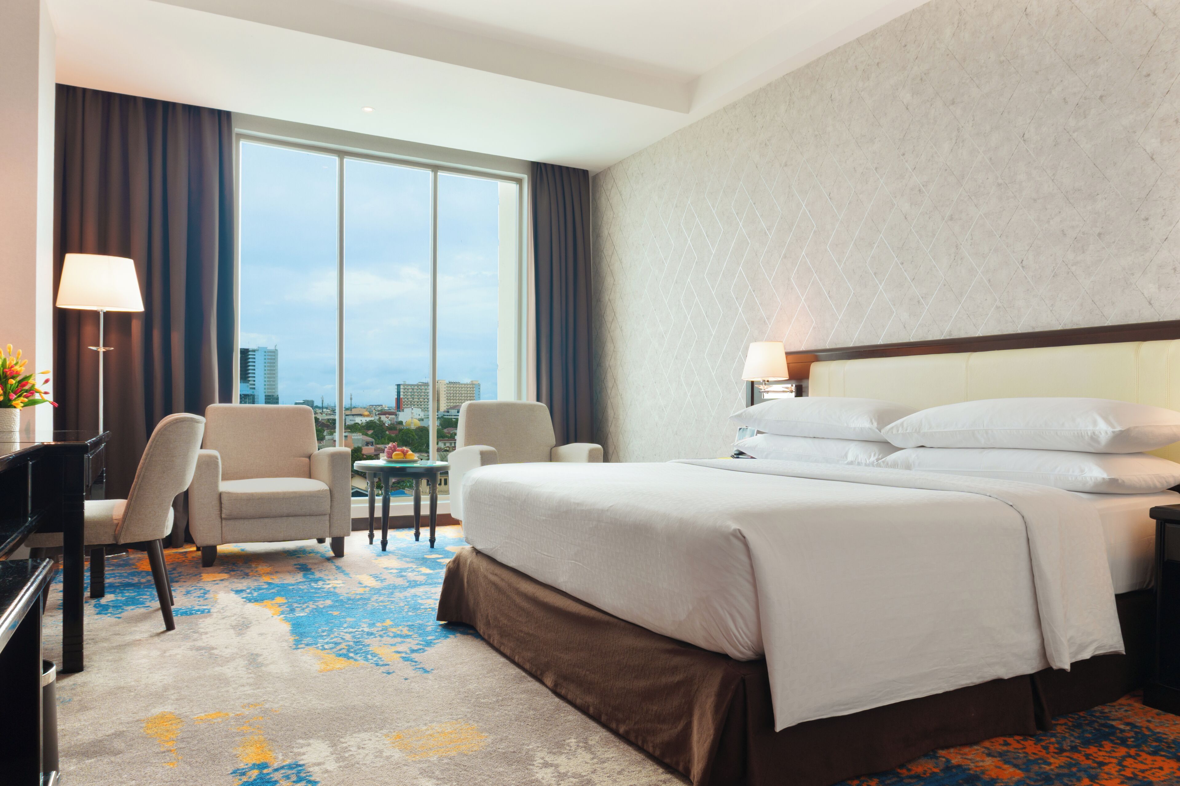 A hotel room with a large bed, two chairs, a desk, a lamp, and a view of the city.