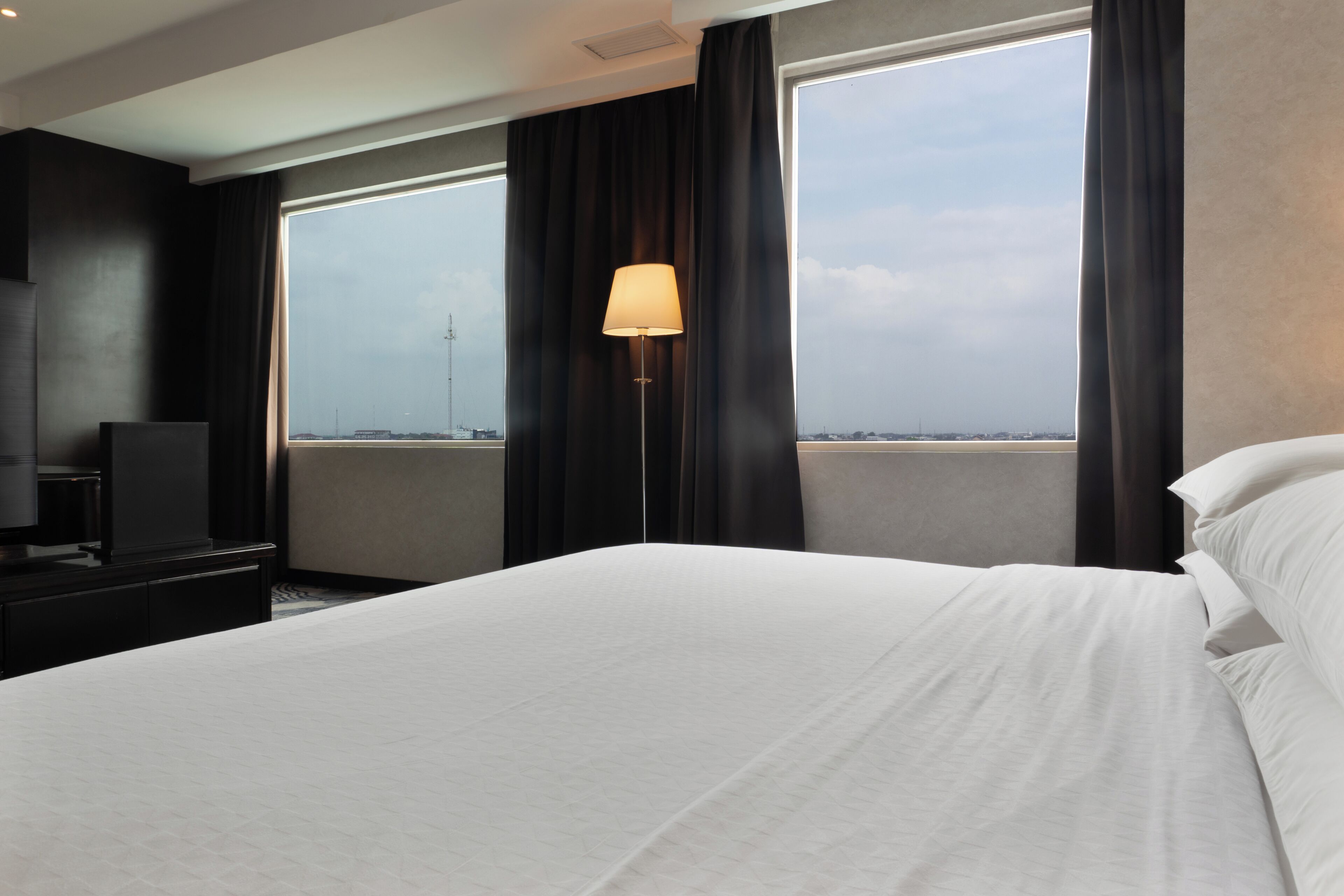 Habitación de hotel con una cama grande, un televisor, una lámpara y vistas de la ciudad a través de amplias ventanas.