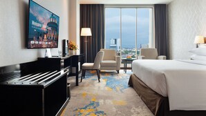 Habitación de hotel con una cama grande, un escritorio con computadora, una silla, una mesita de noche con lámpara y un ventanal con vista a la ciudad.