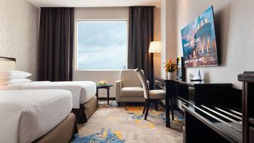 A hotel room with two beds, a desk, a chair, a TV, and a large window.