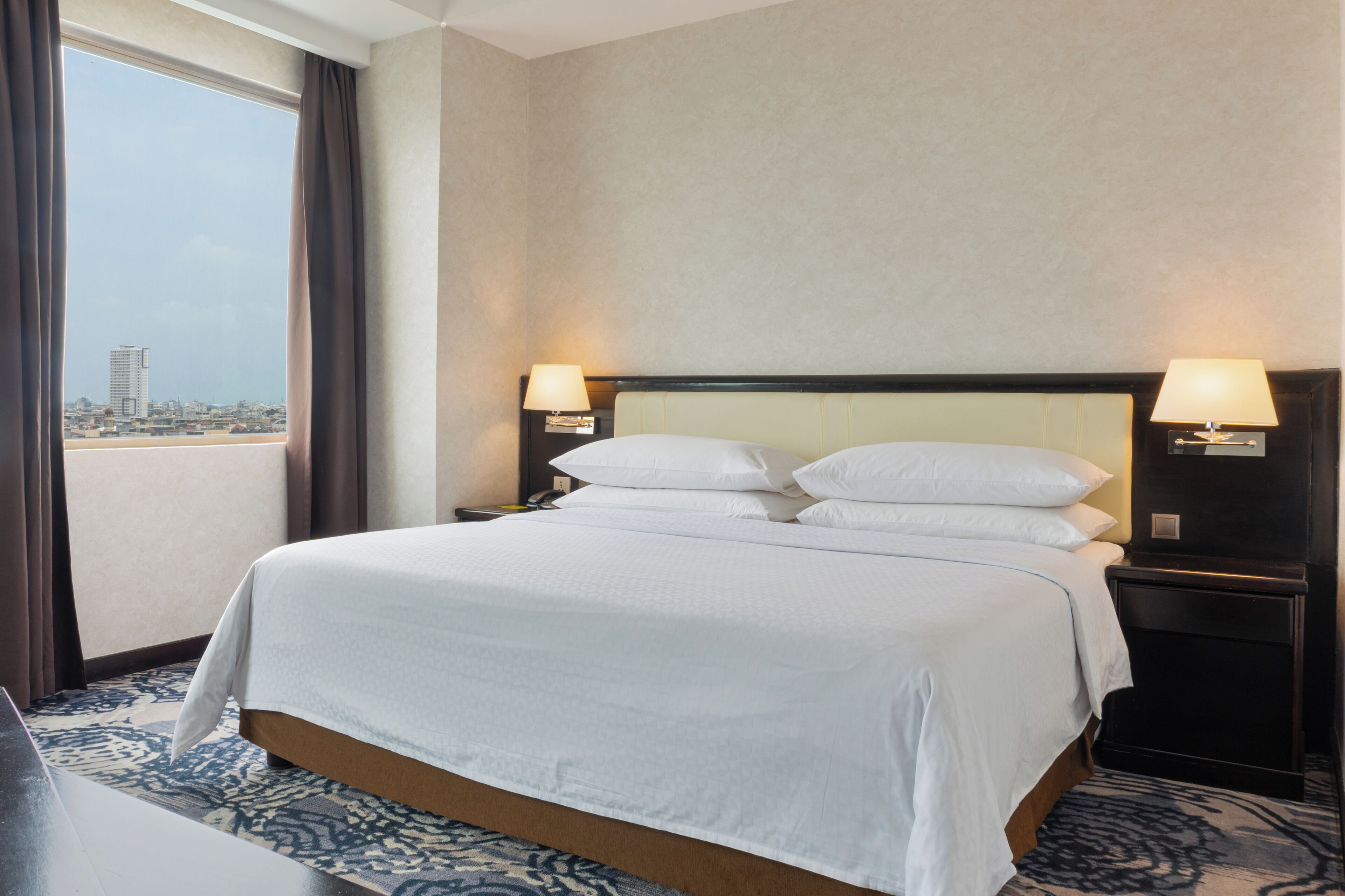Ein Hotelzimmer mit einem großen Bett, zwei Nachttischlampen, Blick auf die Stadt und einem gemusterten Teppich.