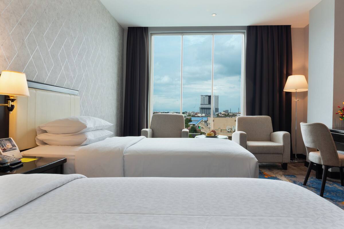 Habitación de hotel con dos camas, un escritorio con lámpara, una silla y vista a la ciudad.
