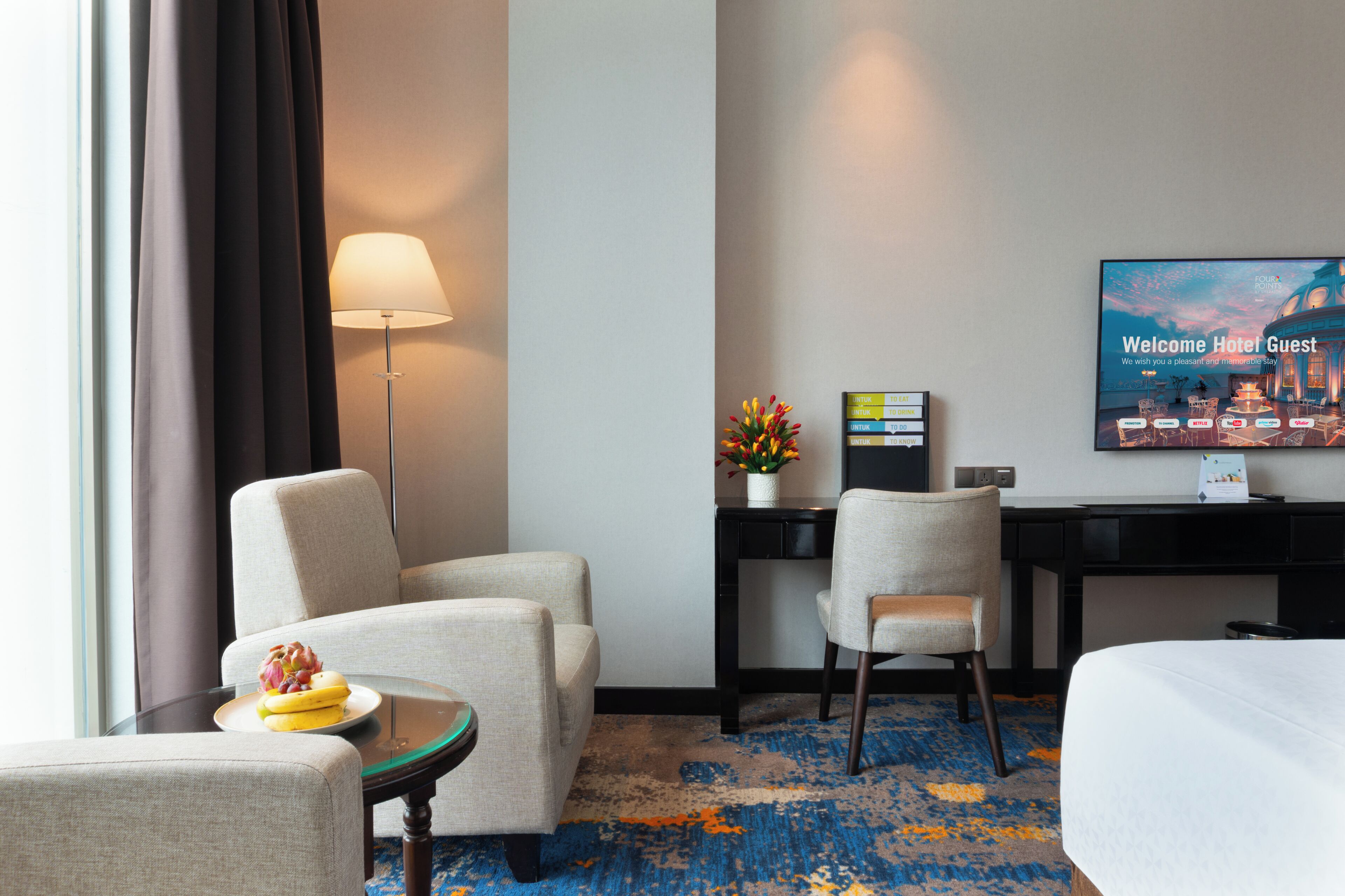 Habitación de hotel con cama, dos sillones, escritorio, lámpara, televisor y alfombra.