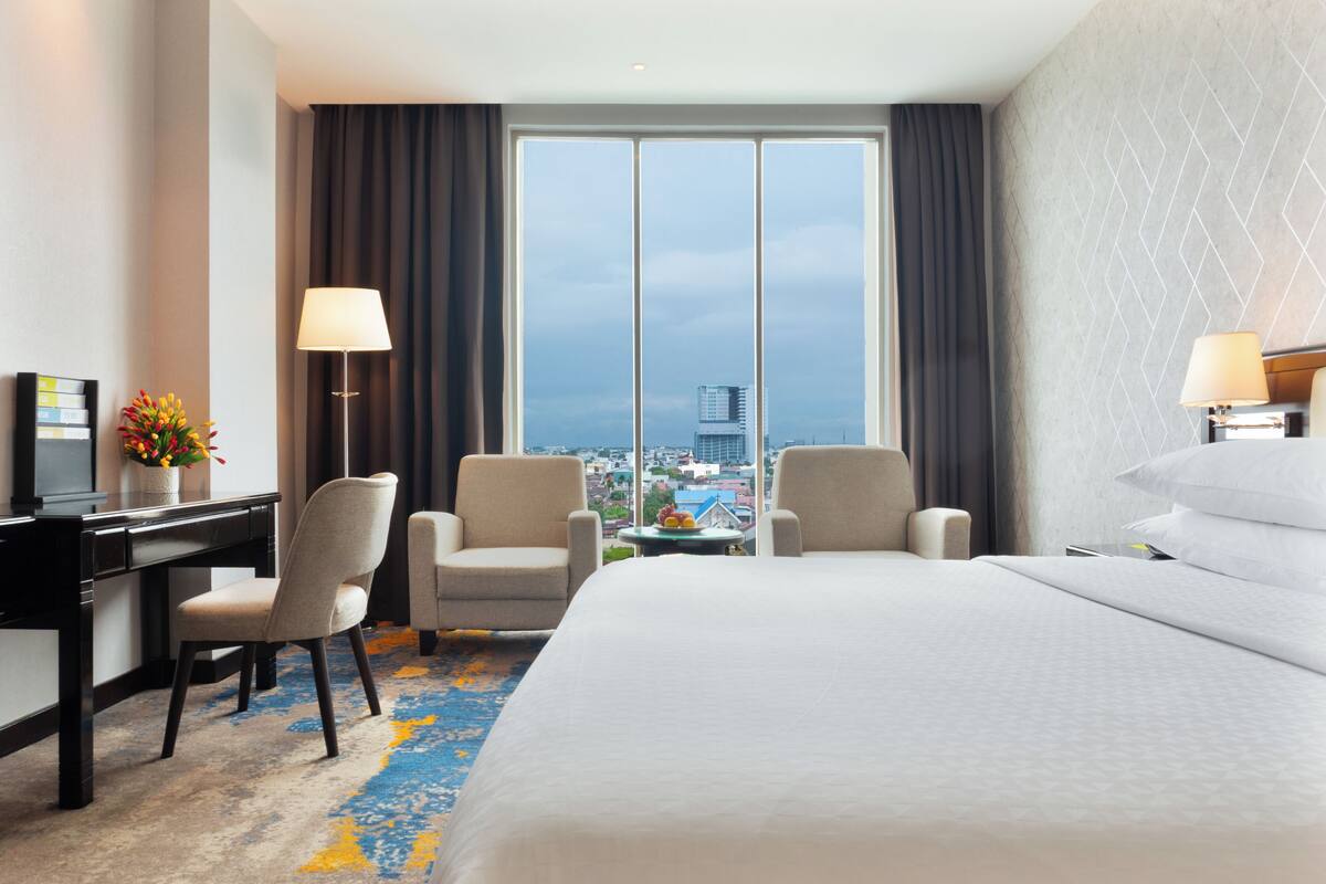Habitación de hotel con cama, dos sillones, un escritorio, una lámpara y vistas a la ciudad.