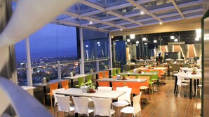 A modern restaurant with high tables and chairs, large windows offering city views, and a mix of green and orange chairs.