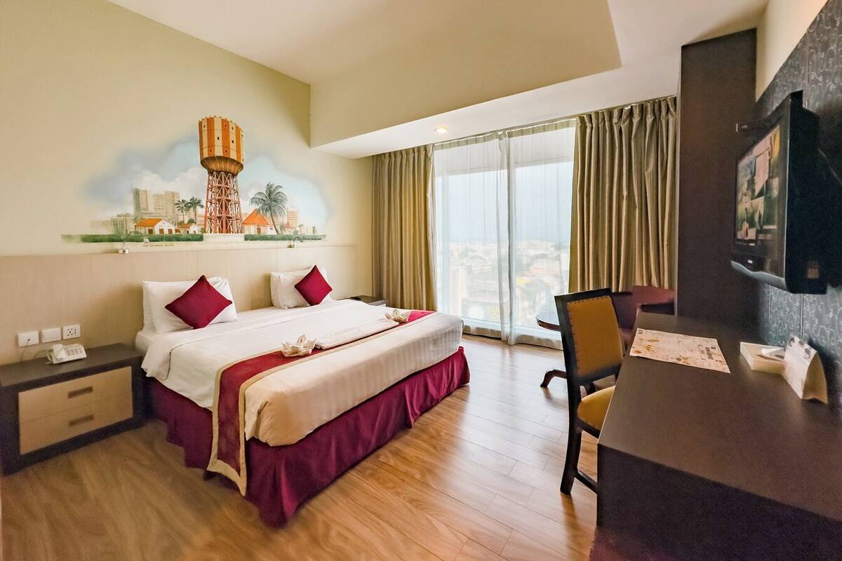 Ein Hotelzimmer mit einem großen Bett, einem Schreibtisch, einem Sessel, einem Fernseher und einem Stadtbild-Wandgemälde.