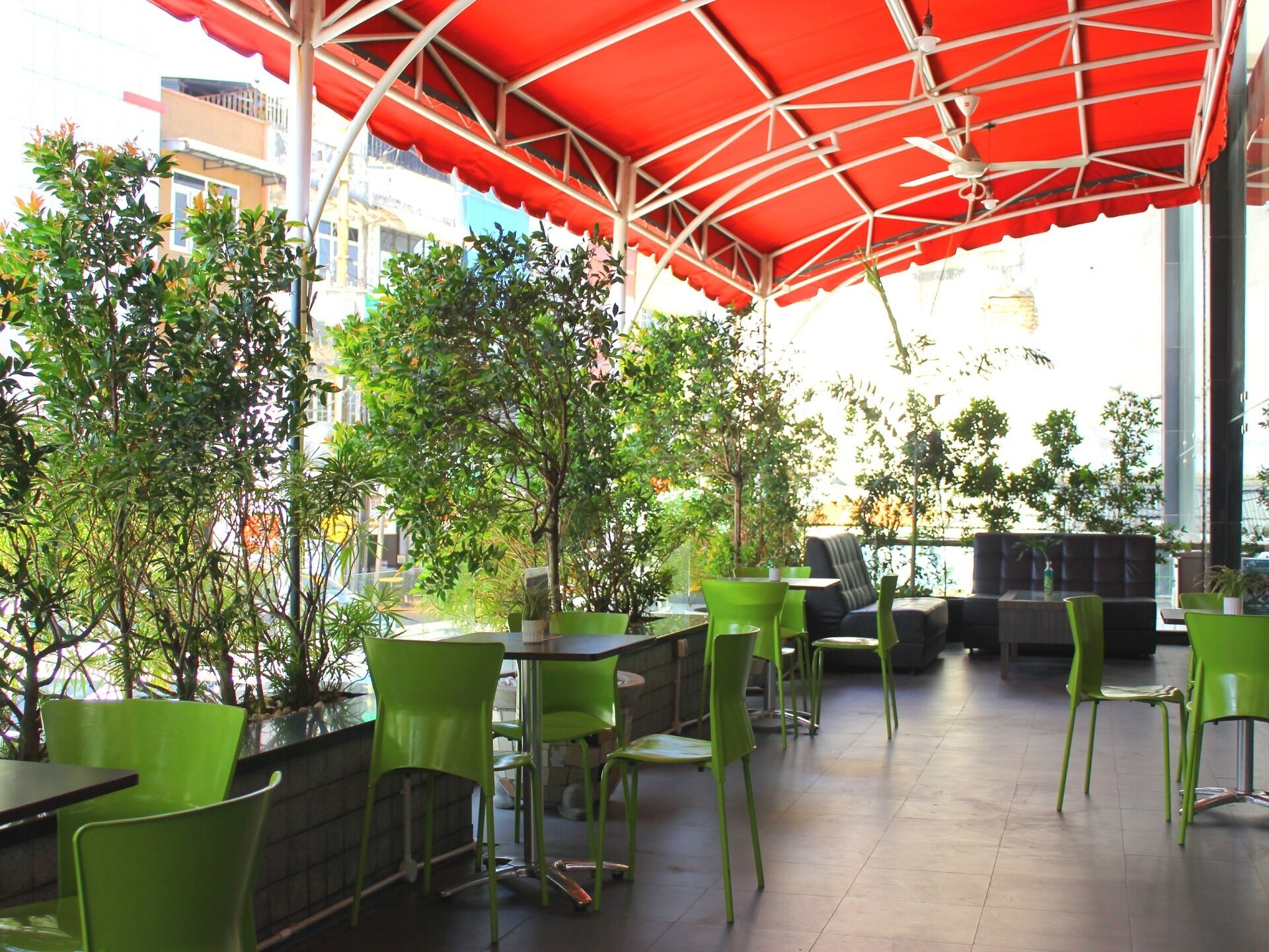 An outdoor seating area with green chairs and tables under a red canopy, surrounded by trees and buildings.