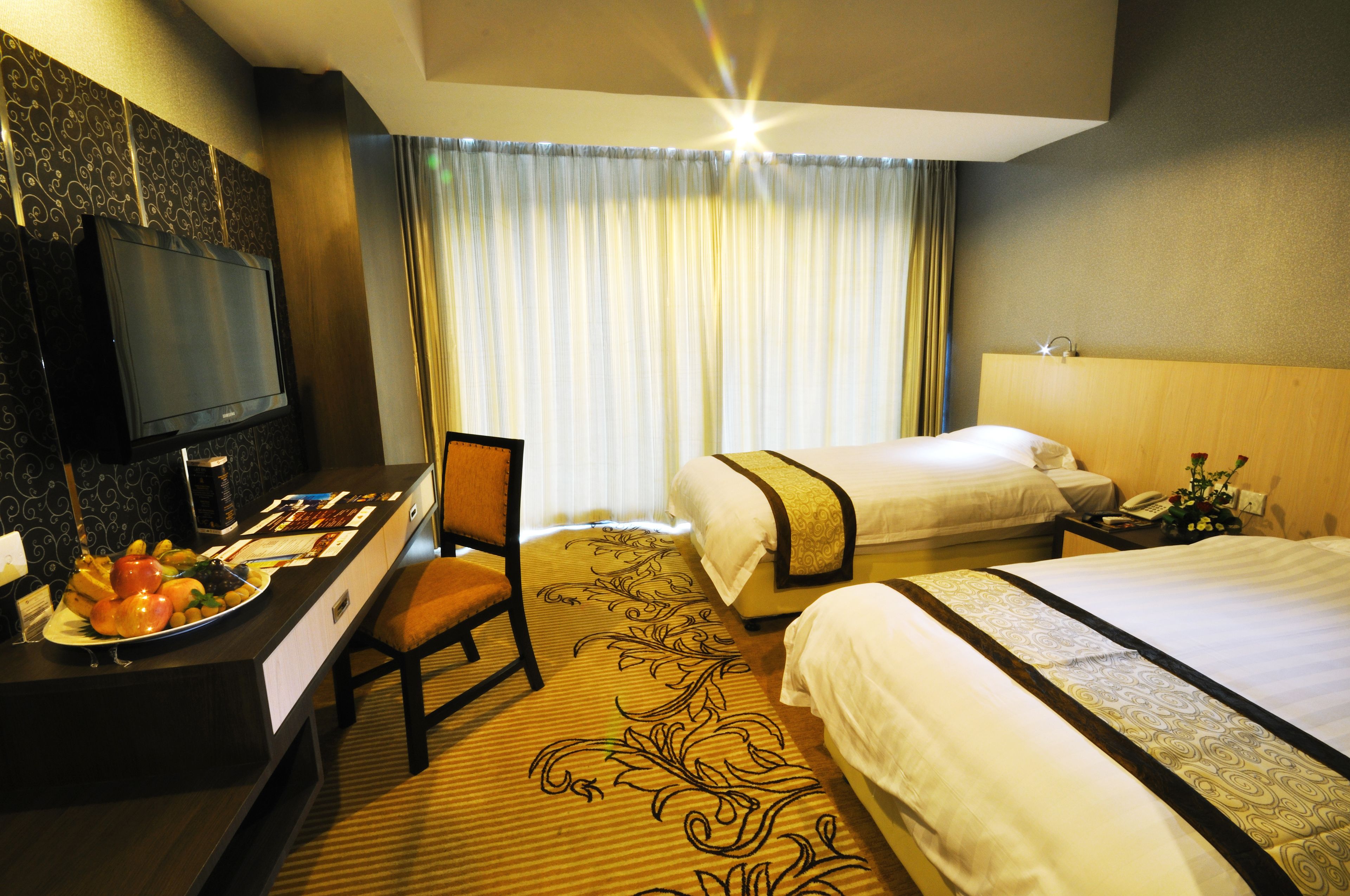 A hotel room with two beds, a desk with a television, and a dining area with a plate of fruit.