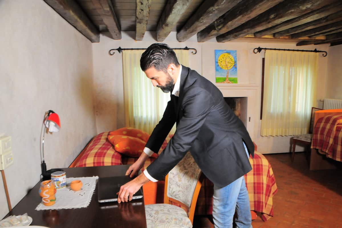 Een man in een pak die een laptop op een houten tafel verzet in een kamer met een open haard, een schilderij en een bed met een geruit plaid.
