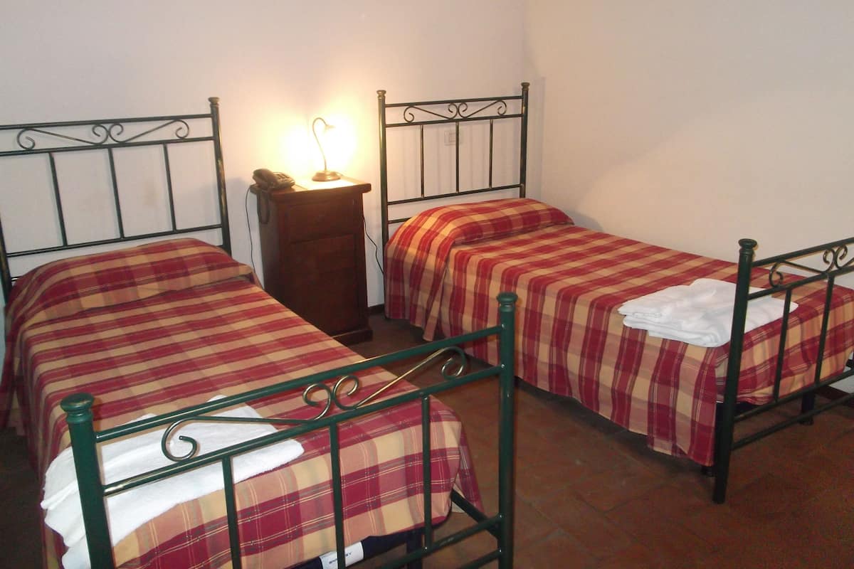 Een kamer met twee eenpersoonsbedden, elk met een metalen frame en ruitbeddengoed. Er staat een houten nachtkastje met een lamp tussen de bedden.