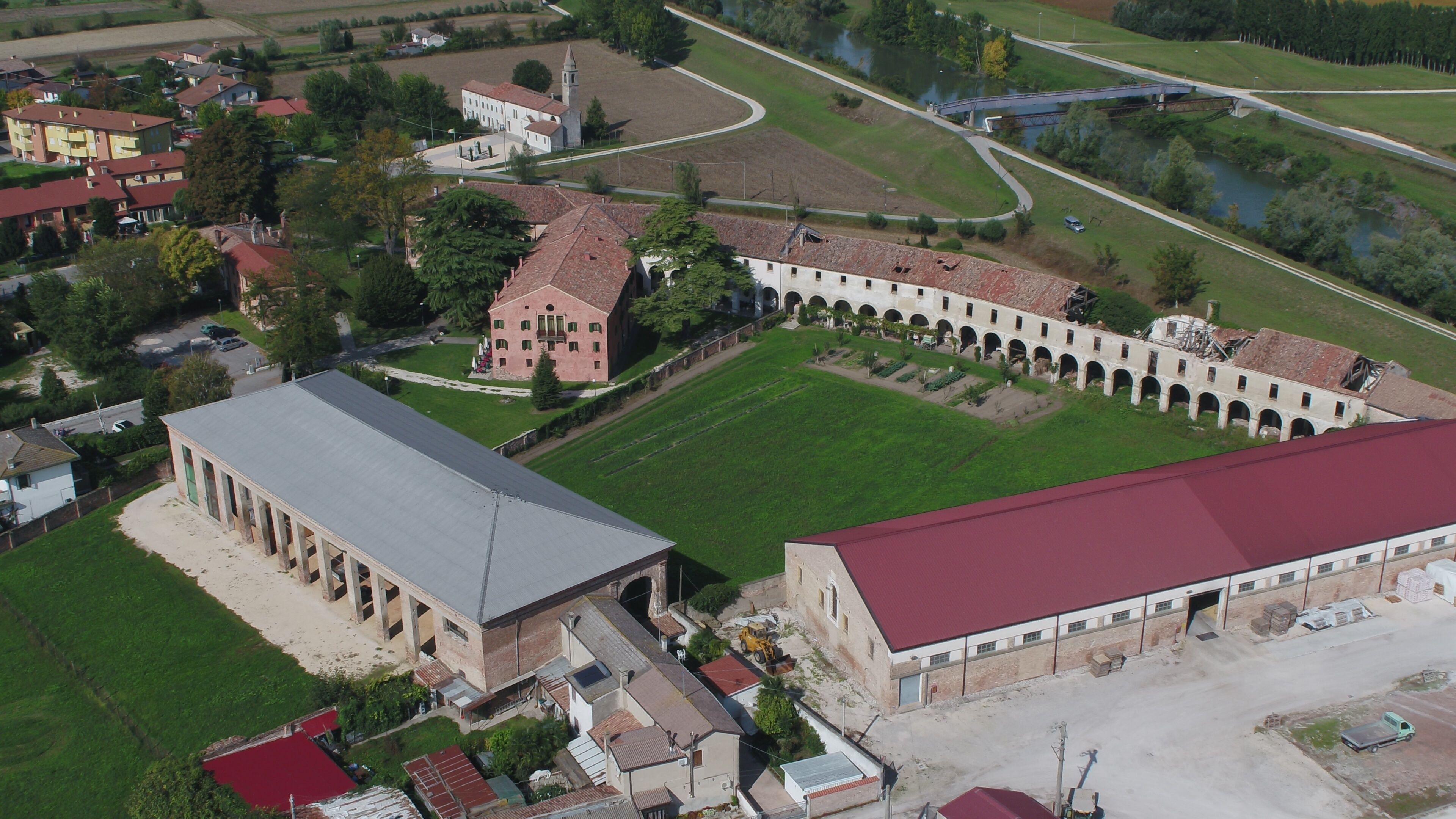 An aerial view of a large building complex with multiple structures, including a central courtyard with arches, a large open building with a flat roof, and surrounding residential buildings.