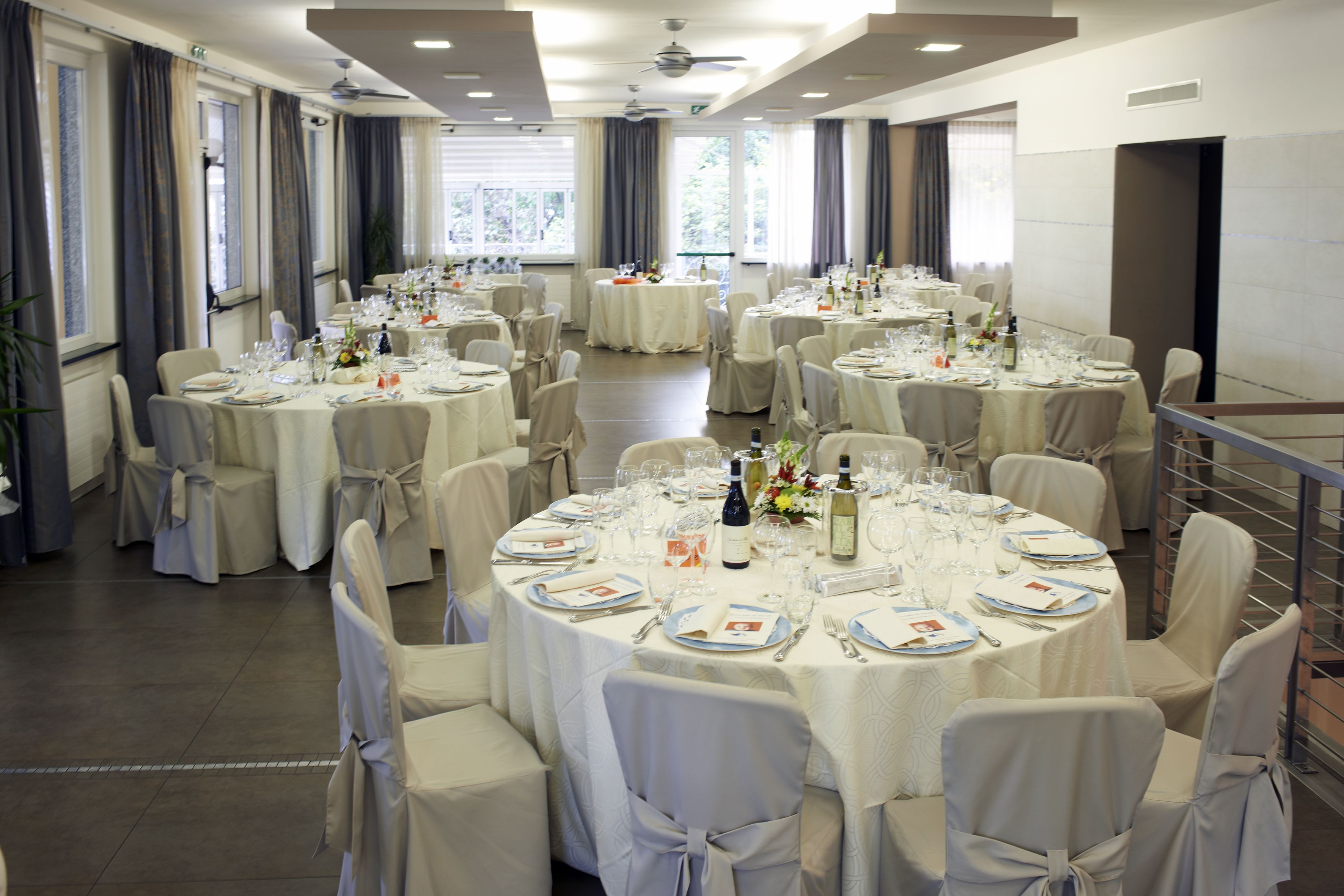 Un salón de banquetes con mesas redondas dispuestas para un evento formal, con manteles blancos y sillas adornadas con lazos.