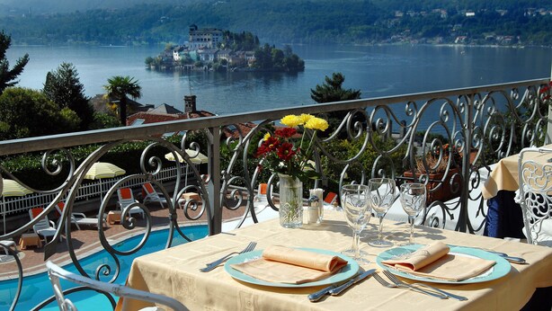 Une table à manger dressée avec une nappe, des assiettes, des couverts et un vase de fleurs, donnant sur un lac et des bâtiments.