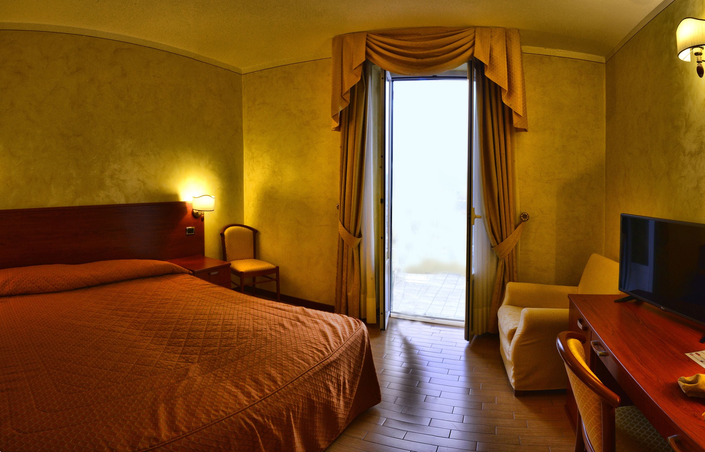 A hotel room with a large bed, a desk with a chair, a television, and a view of the outside through a door.