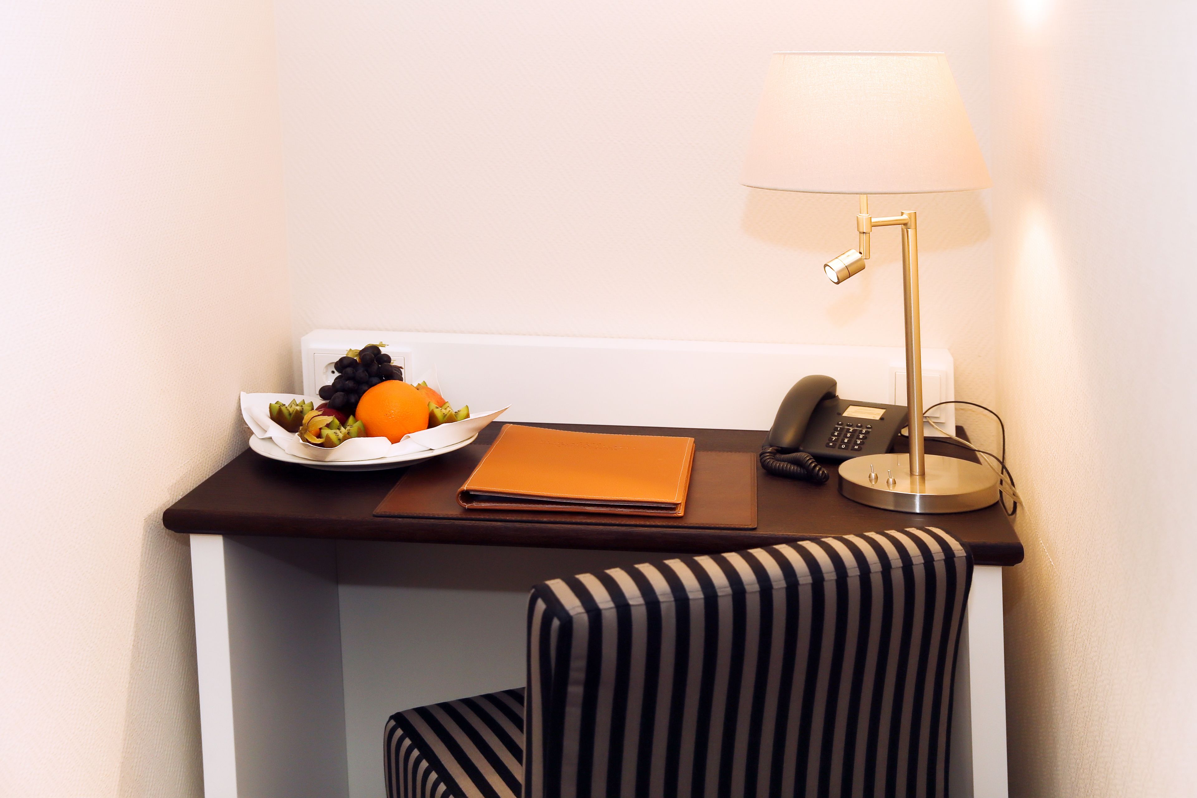Ein Schreibtisch im Hotelzimmer mit einer Obstplatte, einer Lampe, einem Telefon und einem gestreiften Stuhl.