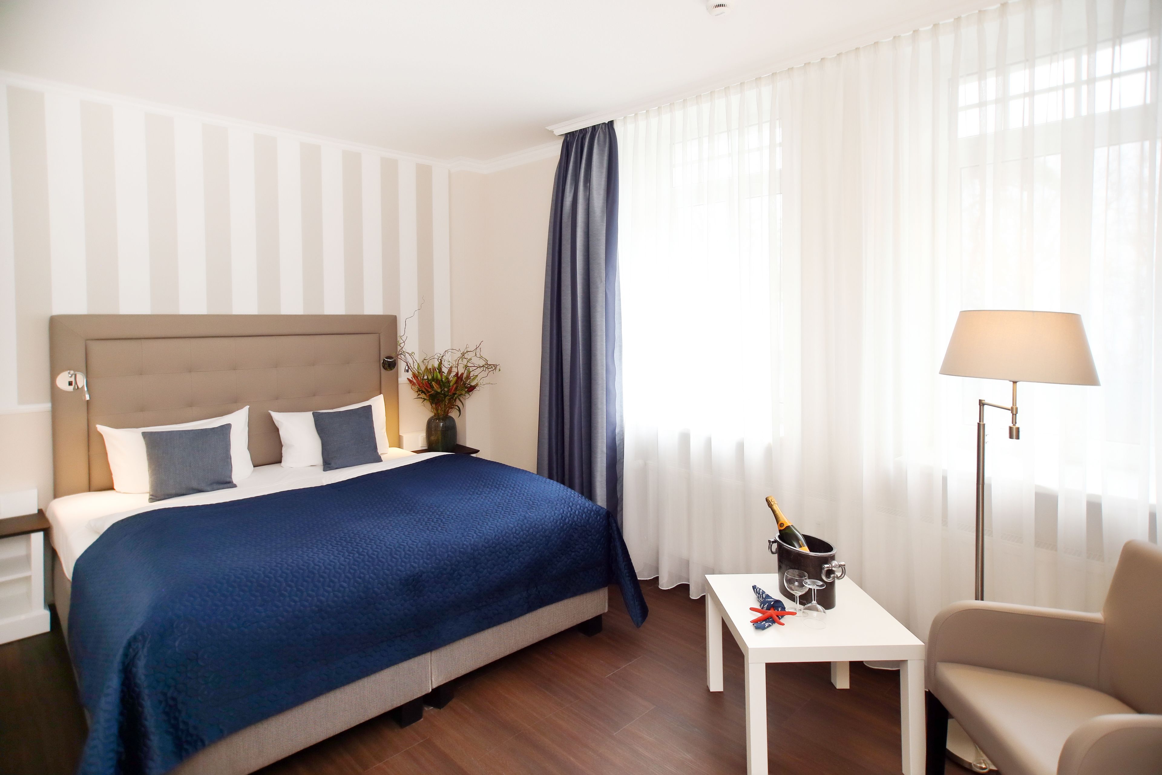 A hotel room with a bed, a nightstand, a bedside table with a lamp, a chair, and a window with curtains.