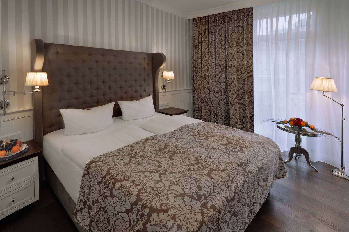 Una cama bien hecha con una colcha estampada, dos lámparas de noche, una mesita con un florero y una mesita de noche con un frutero.