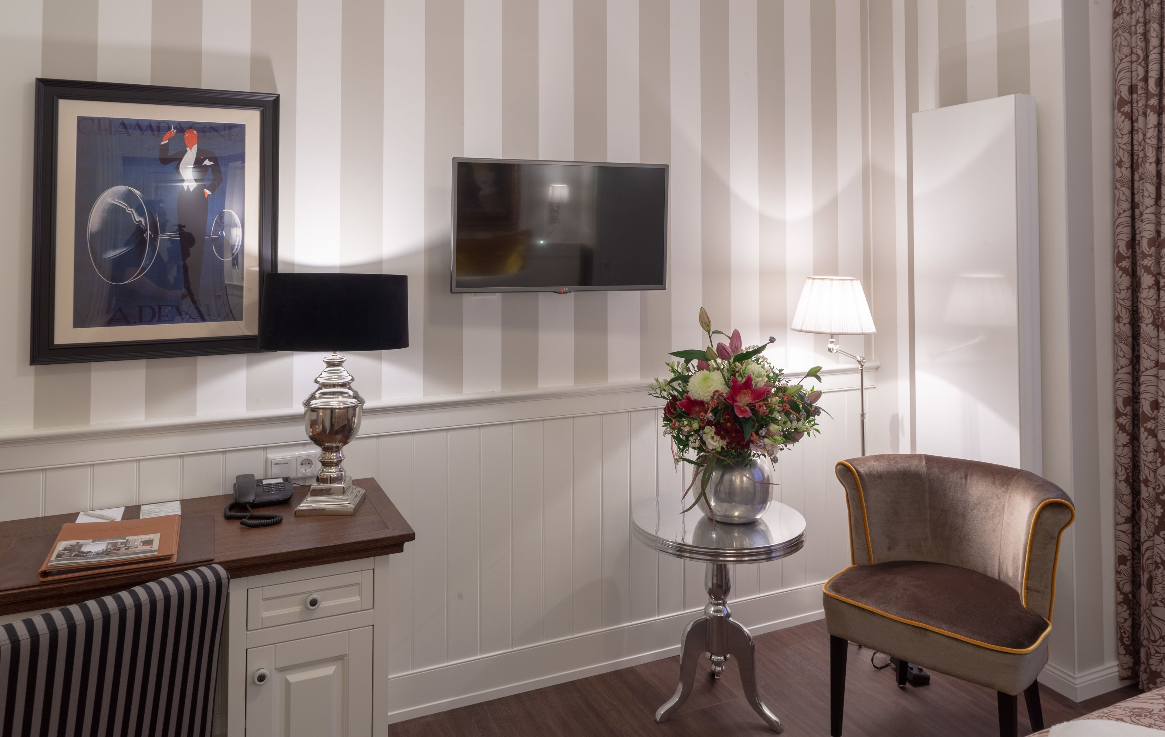 Et hotelværelse med et fjernsyn ophængt på væggen, et skrivebord med en telefon, en vase med blomster og en stol.