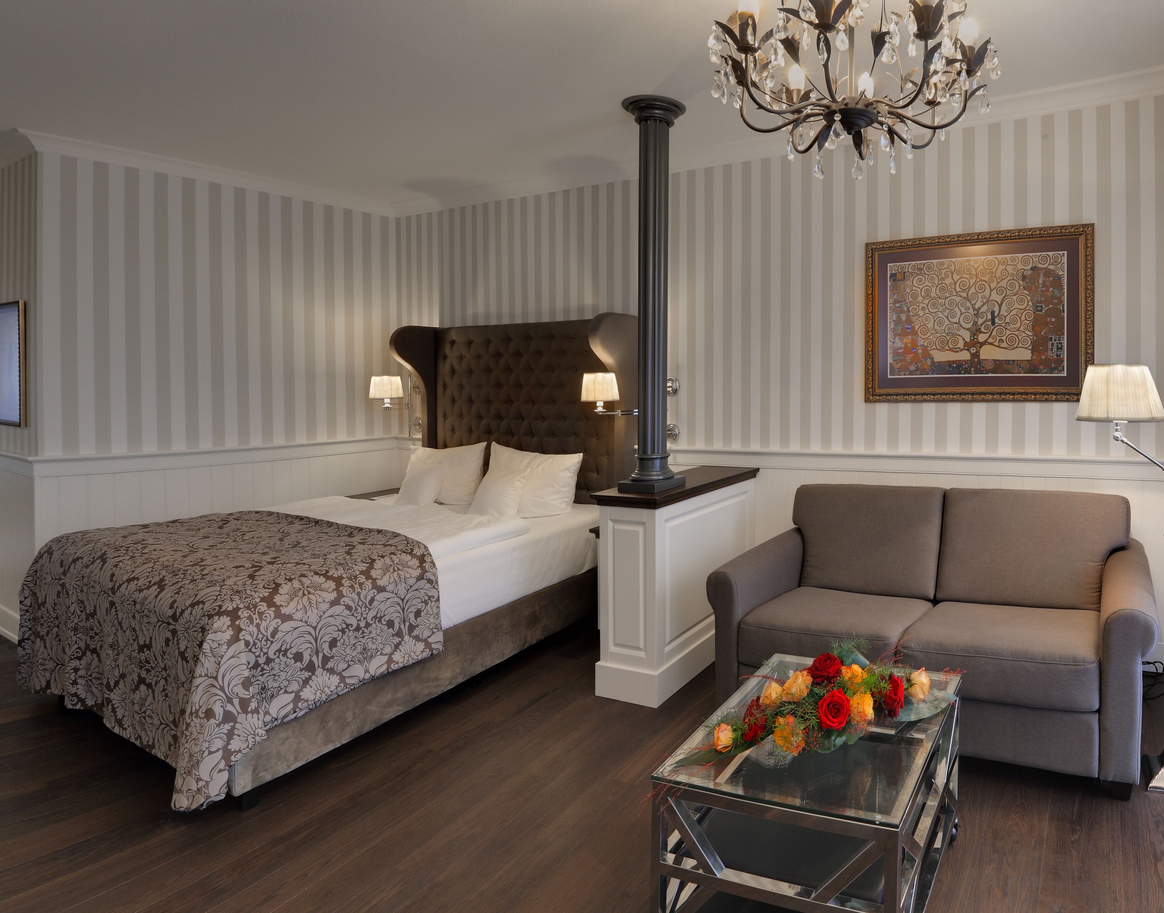 Un cuarto de hotel con una cama grande, un sofá, una mesa de centro de vidrio con flores, un candelabro y un cuadro en la pared.