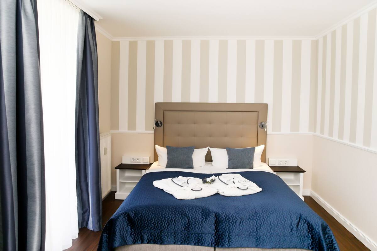 Una cama bien hecha con un edredón azul, dos almohadas y un cabecero. Hay dos mesitas de noche con lámparas, una ventana con cortinas y una pared a rayas.